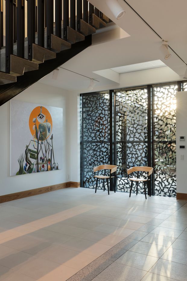 A bright contemporary interior lobby or circulation space with a dramatic open stair overhead. The stair has dark metal stringers and vertical balustrade elements with projecting timber or bronze-toned treads. Pale tiled flooring, white walls, timber skirting, recessed and linear ceiling lighting, and a large rectangular skylight create an airy architectural setting. Full-height black-framed glazing is screened with an intricate patterned privacy treatment, with two timber-and-black chairs positioned in front. A colourful artwork is mounted on the left wall.