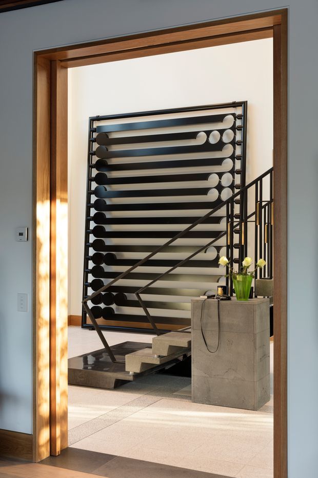 A contemporary interior view through a timber-framed doorway into a stair hall. The space features a sculptural floating staircase with dark metal railings, a large geometric black-and-white decorative screen or partition behind it, pale tiled flooring, and a substantial concrete pedestal with a vase of flowers. The composition is architectural and refined, with strong linear forms and warm natural light.