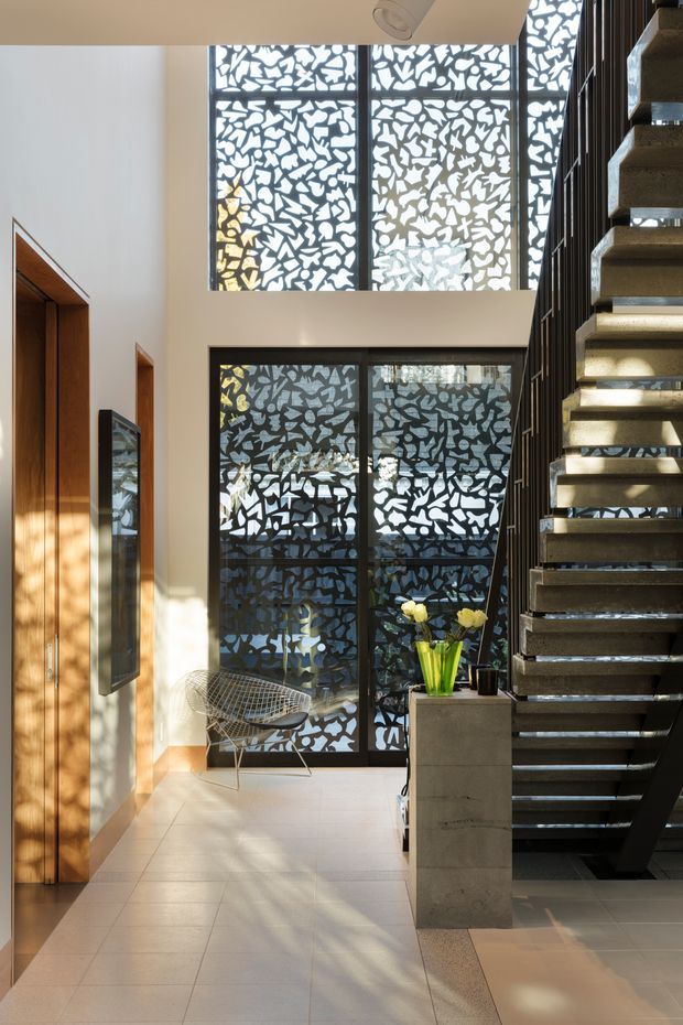 A bright, double-height residential entry hall with a floating open-tread staircase and dark metal balustrade on the right. Large black-framed windows feature distinctive patterned privacy screens, while warm timber doors and panelled joinery line the left wall. Pale tiled flooring, white walls, a sculptural wire chair, and a small concrete planter with flowers complete the contemporary interior.