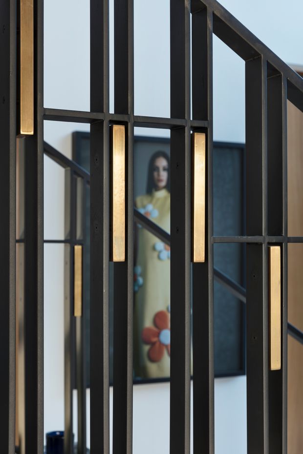 Close-up interior view of a dark metal framed decorative screen or balustrade with repeated rectangular geometric divisions and integrated warm linear lighting inserts. A staircase and framed artwork are softly visible behind the screen, creating a layered architectural composition.