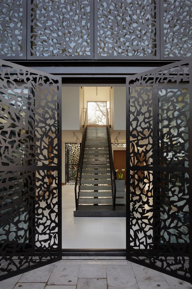 View through open decorative metal entry gates into a modern interior stair hall. Intricately perforated dark screens frame a broad staircase with dark treads and slim metal balustrades, leading toward a bright window. Pale walls and flooring contrast with the black patterned screens, while additional matching screens and a timber door are visible inside.