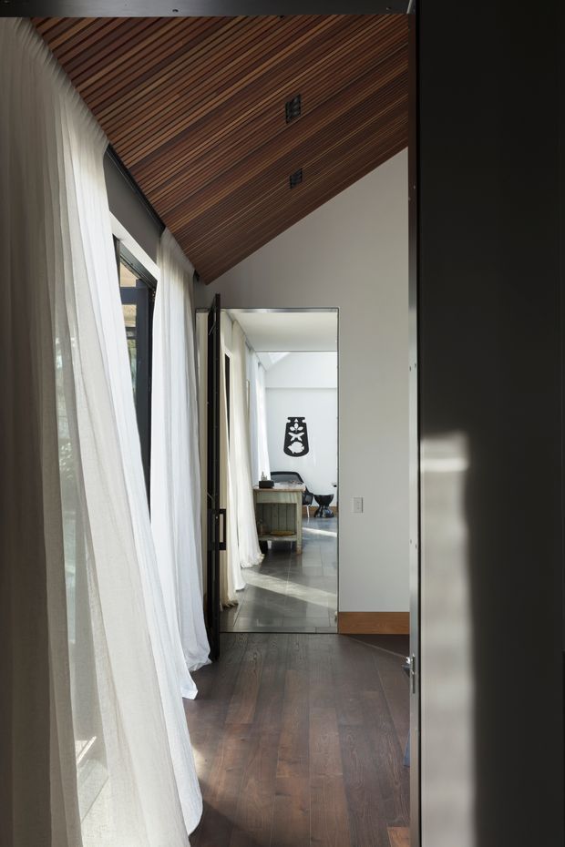 A narrow, softly lit interior passage with dark timber flooring, sheer white curtains along tall glazing, white walls, and a dramatic sloped ceiling lined with narrow warm-toned timber boards. A full-height mirror or glazed opening reflects a brighter adjoining area with furniture and additional glazing. Dark-framed openings and restrained detailing create a calm, contemporary architectural interior.