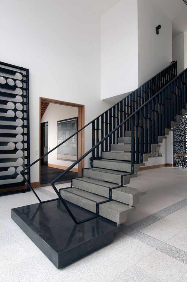A dramatic interior stairway with floating grey concrete steps, slim black metal balustrades, and a polished black stone plinth. The stair rises through a bright, high-ceilinged foyer, with white walls, pale terrazzo-like flooring, timber-framed openings, and a bold black-and-white decorative wall feature.