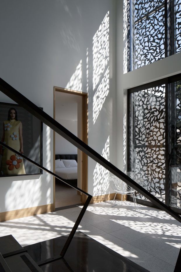 A bright double-height interior landing or hallway with a dramatic black stair railing in the foreground. White walls and pale tiled flooring are patterned with strong dappled sunlight cast through decorative perforated metal screens beside tall windows. A bedroom is visible through an open doorway, while a framed artwork and a white wire chair add sparse furnishings. The space has a clean, contemporary architectural character with strong geometric contrast and sculptural light.