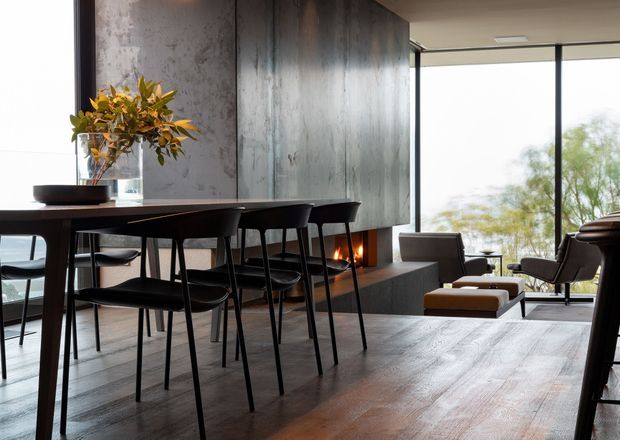 A refined open-plan dining and living area with a dark timber dining table and matching black chairs in the foreground. A textured grey feature wall incorporates a linear fireplace, while the lounge area beyond has leather seating and a large glazed opening overlooking trees. The space uses dark timber flooring, muted grey finishes, and warm firelight for a sophisticated, contemporary atmosphere.
