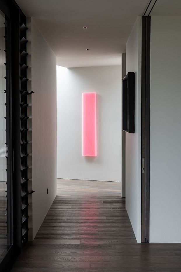 A minimalist interior hallway with pale plaster walls, dark timber flooring, black-framed glazing or cabinetry, and a vivid vertical pink illuminated wall feature at the end of the corridor. Recessed ceiling lights and concealed doors create a clean, refined architectural composition.