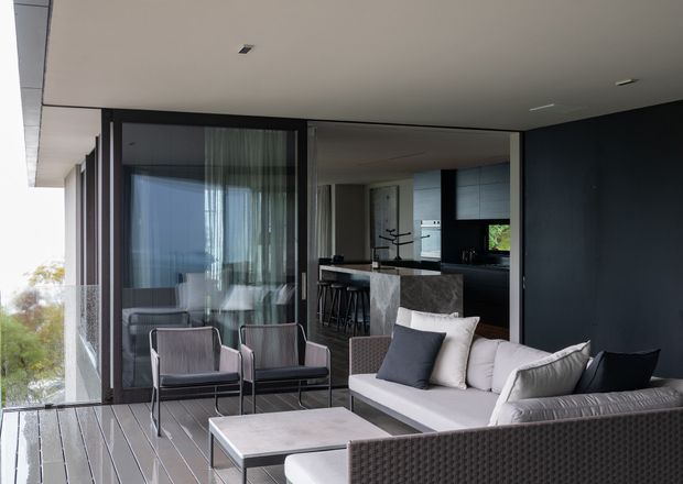 Contemporary covered outdoor terrace or balcony adjoining an open-plan interior, furnished with a large pale sectional sofa, woven armchairs, and a low rectangular coffee table. Full-height glazing, dark-framed sliding doors, dark wall panels, and a compact bar or kitchen area create a refined indoor-outdoor setting. Timber decking and abundant natural light are prominent.