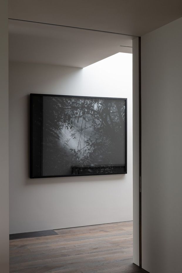 A minimalist interior passage or gallery-like space viewed through a partially open doorway. White walls and a clean recessed ceiling line frame a large dark wall-mounted artwork or screen, while wide-plank timber flooring adds warmth. The space is sparsely furnished, with a strong diagonal opening of bright light creating a quiet, refined atmosphere.