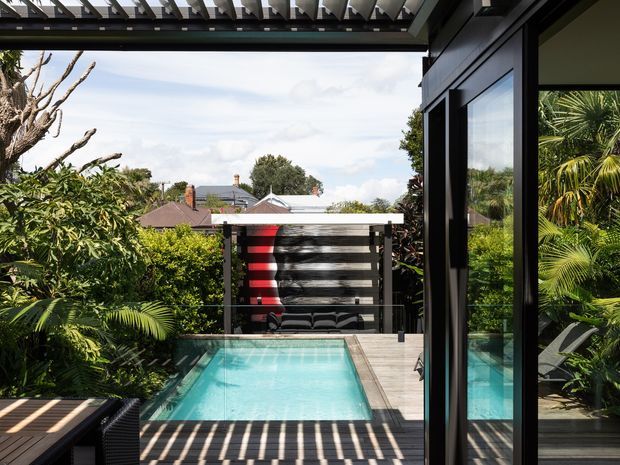 A compact outdoor swimming pool viewed from a dark-framed glazed interior or covered terrace. The pool is surrounded by dense tropical planting and timber decking, with a slatted privacy screen and a bold red-and-black outdoor feature beyond. Overhead pergola slats cast strong linear shadows across the deck, while neighbouring rooftops are visible in the distance.