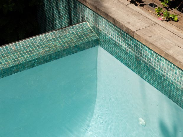 Close-up view of a swimming pool corner with turquoise mosaic-tiled walls, a submerged tiled bench or step, pale blue pool water, and a broad natural stone coping edge. Vegetation is visible around the perimeter.