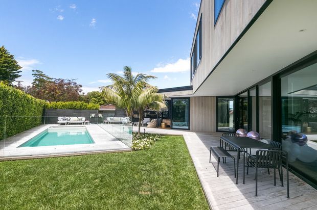 Exterior view of a contemporary two-storey residence opening onto a landscaped backyard with a rectangular swimming pool, glass pool fencing, timber-look decking, and a covered outdoor dining area. The home features strong horizontal lines, extensive dark-framed glazing, pale vertical cladding, and a flat roof. Palm and hedge planting create a bright resort-like setting.