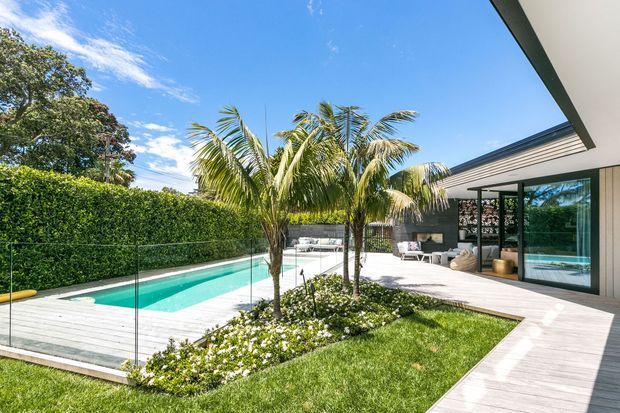 A bright contemporary courtyard-style outdoor area featuring a rectangular swimming pool enclosed by clear glass balustrading, a pale timber deck, tropical palm trees, clipped hedging, and a modern house with broad glazed openings and a low-pitched roof. The space includes a poolside lounge area and a walkway connecting the lawn to the house.