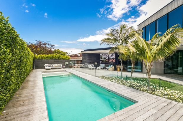 Bright contemporary residential exterior featuring a long rectangular swimming pool surrounded by pale timber decking, a landscaped courtyard with palms and clipped hedges, and a modern dark-clad home with expansive blue-tinted glazing and a flat roof.