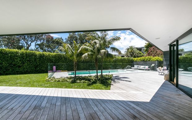 A modern covered outdoor living area opens onto a broad timber deck and landscaped backyard with a narrow swimming pool, palm trees, clipped hedges, and outdoor lounge furniture. The image emphasizes the seamless indoor-outdoor connection, large glazed openings, and a dramatic angular roof overhang.