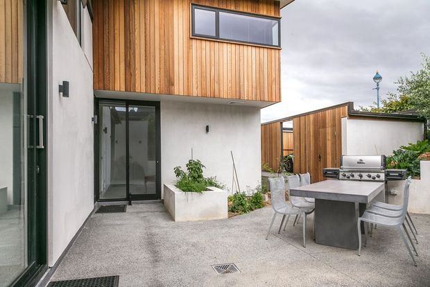 A contemporary outdoor courtyard and dining area beside a multi-level home. The building combines warm vertical timber cladding with smooth white rendered walls and dark-framed glazing. A built-in outdoor kitchen with stainless-steel barbecue sits beside a concrete dining table and modern chairs on a textured paved surface, with raised planters adding greenery.