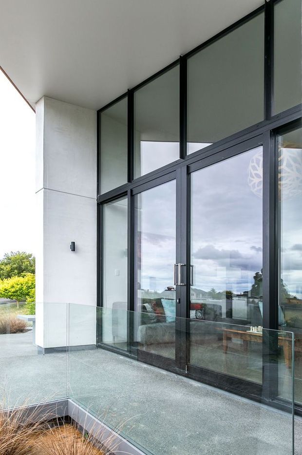 Contemporary exterior terrace or balcony with expansive black-framed floor-to-ceiling glazing and glazed double doors. Pale rendered wall panels, a glass balustrade, and a grey textured deck create a clean modern facade, with the interior and cloudy landscape reflected in the glazing.