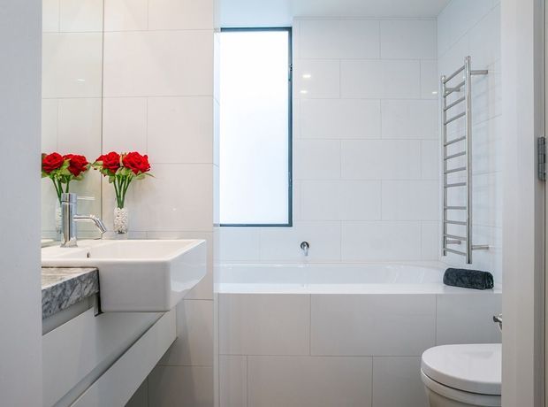 Bright, clean contemporary bathroom with a white tiled bath surround, built-in bathtub, chrome heated towel rail, wall-mounted shower fitting, floating vanity, stone-look benchtop, and white toilet. Red flowers provide a small decorative accent.