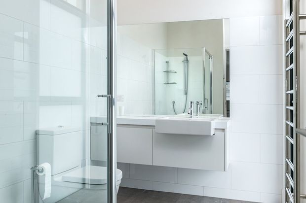 Bright contemporary bathroom featuring white rectangular wall tiles, a wall-mounted white vanity with integrated basin, large mirror, glass-enclosed shower, toilet, chrome tapware and a vertical heated towel rail. The palette is predominantly white with grey stone-look flooring and chrome accents.