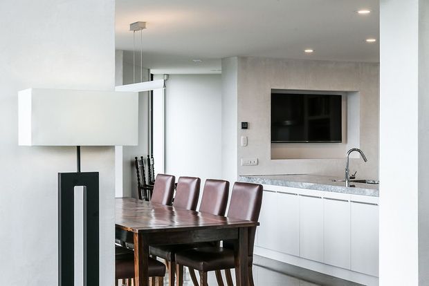 Modern open-plan dining and kitchen interior with a dark wood dining table and upholstered dining chairs in the foreground. The white kitchen features handleless cabinetry, a pale stone benchtop, stainless steel mixer tap, and a grey textured feature wall with an integrated television. A minimalist pendant light and recessed ceiling lights complete the clean, contemporary space.