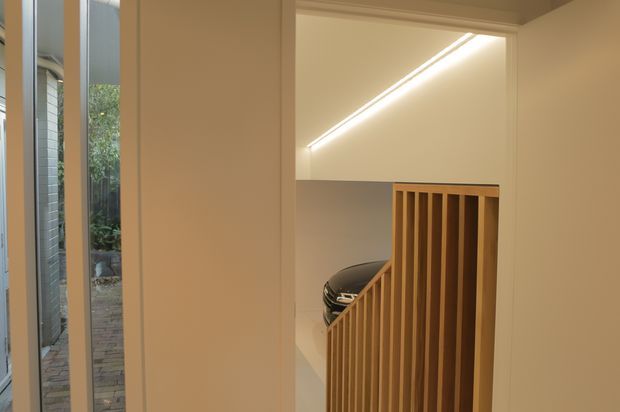 View from an interior hallway toward a modern stairwell and garage area. A warm timber slatted stair balustrade contrasts with smooth off-white walls and ceiling, while a linear recessed light illuminates the stair void. A car is partially visible below, and glazing at left looks onto a narrow landscaped passage.