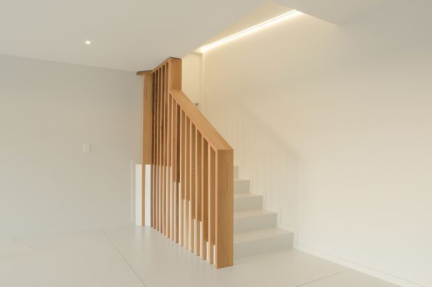 Minimalist interior view of a stairway with pale timber vertical balustrade slats and handrail, white painted walls, light flooring, and recessed linear ceiling lighting. The staircase rises along the wall, creating a clean, bright and understated space.
