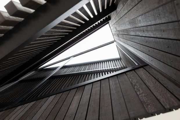 Upward-looking architectural view through a tall central void, framed by dark vertical timber-like wall cladding and black metal slatted balconies or walkways. Repeated horizontal and vertical lines create a dramatic geometric composition around the bright open sky.