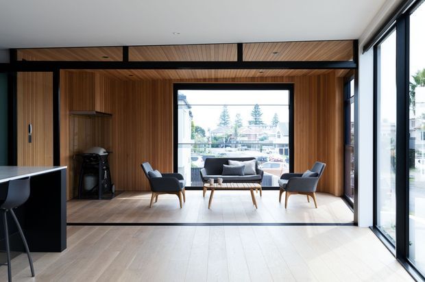 A bright, contemporary open-plan living area with extensive floor-to-ceiling glazing and a wide view toward an outdoor deck or courtyard. The room features warm timber wall and ceiling panelling, pale timber flooring, a dark built-in kitchen or island element at left, and a mid-century-inspired seating arrangement with two lounge chairs, a sofa, and a timber coffee table. Black-framed glazing and structural detailing provide contrast.