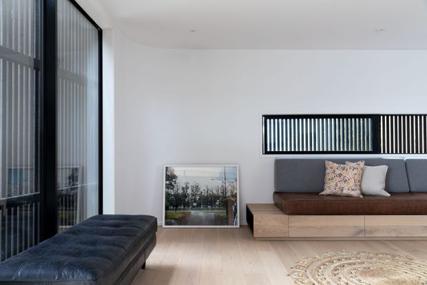 Minimal contemporary living room with a low built-in timber media or seating unit, dark grey upholstered bench seating in the foreground, a grey sofa with patterned cushions, pale timber flooring, and large black-framed glazed openings with vertical screening. The room is bright and understated, with a framed artwork leaning against the wall and a woven rug partially visible.