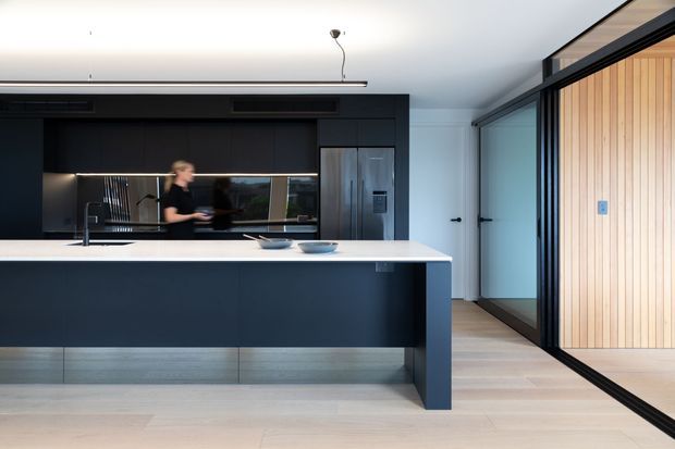 Contemporary kitchen with a large white island and dark matte cabinetry, integrated stainless-steel appliances, pale timber flooring, and a black-framed glazed partition beside a vertical timber-lined room. The space is clean-lined and minimally styled, with a restrained black, white, grey, and light-wood palette.