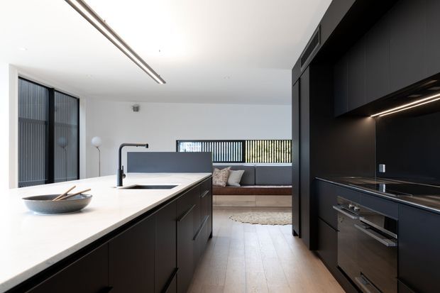 A contemporary open-plan kitchen viewed from the island, featuring a large white stone benchtop, dark matte cabinetry, integrated appliances, black tapware, linear pendant lighting, and timber flooring. Beyond the kitchen is a compact living area with a built-in bench seat and vertical screened glazing.