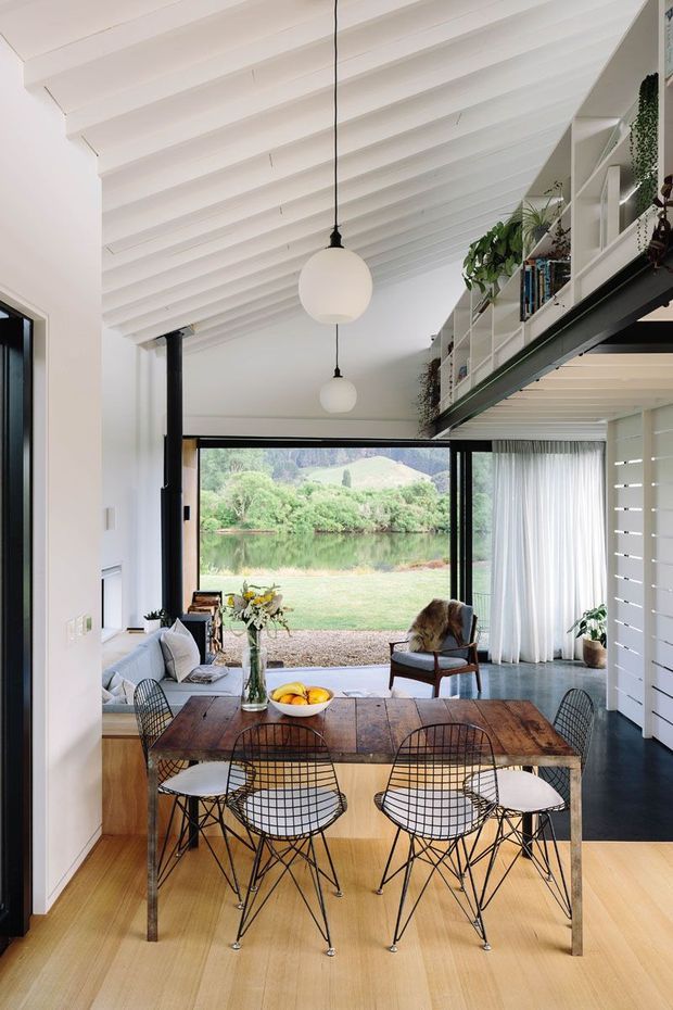 A bright open-plan dining and living space with a long timber dining table and black wire-frame chairs in the foreground. The room has a dramatic white pitched ceiling with exposed painted rafters, black structural posts and beams, pendant globe lights, pale timber flooring, and extensive glazing opening toward a landscaped waterside view. Built-in white shelving and trailing plants occupy a mezzanine-like upper level along the right side, while a sofa and lounge chair form the living area beyond the dining zone.