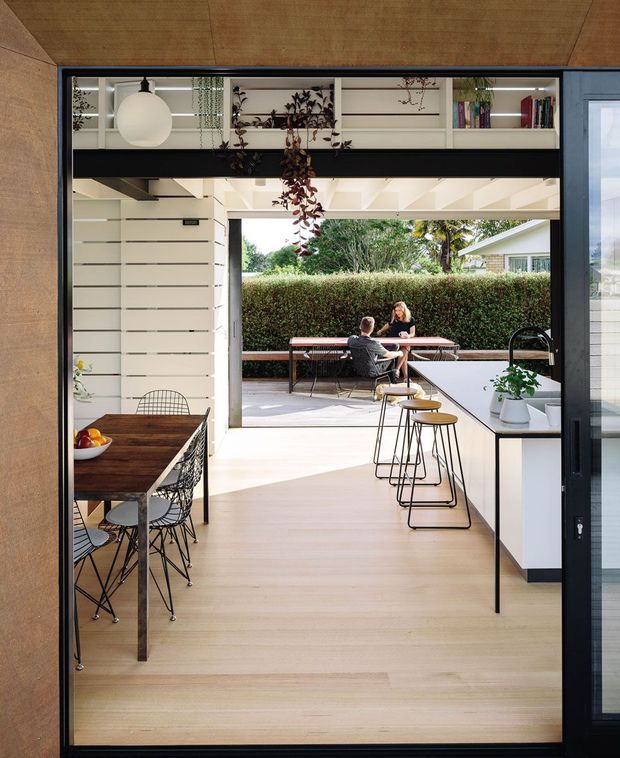 A bright, open-plan kitchen and dining area viewed through a large glazed opening toward an outdoor terrace. The interior features pale timber flooring and ceiling lining, white horizontal wall cladding, white built-in shelving with integrated lighting and books, dark structural beams and window frames, a dark timber dining table with wire chairs, and a white kitchen island with slim black stools. The terrace beyond includes a long dining table and bench seating, with hedging screening the garden. The space has a clean contemporary character with strong indoor-outdoor connection.