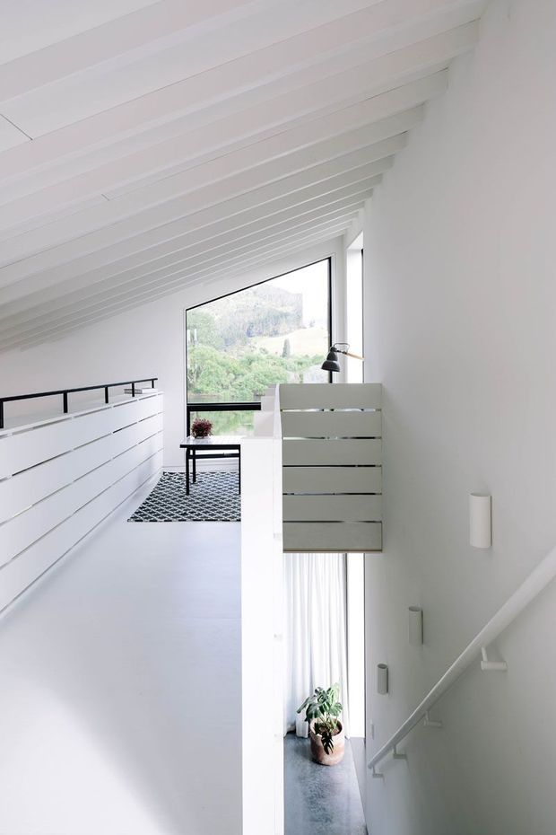 Bright minimalist interior view of an upper-level landing or hallway beside a stairwell. The space has white walls, white-painted exposed ceiling beams, a white balustrade, black metal handrails, and a suspended or built-in white slatted cabinet. Large windows provide views of a green landscape, while a narrow stair descends to a lower level with sheer curtains and a potted plant. A small black table, lamp, and patterned rug furnish the landing.