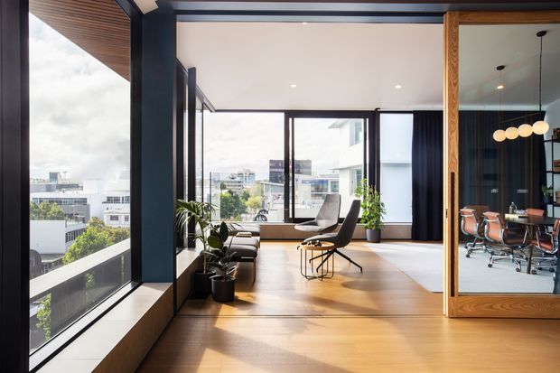 A bright contemporary office or studio interior with expansive floor-to-ceiling corner glazing overlooking an urban setting. The main space features warm timber flooring, dark structural framing, a low lounge seating area, potted plants, and a sculptural lounge chair. Through a large timber-framed glass partition, a meeting room is visible with a conference table, office chairs, pendant globe lighting, and dark curtains. The ceiling has exposed dark beams and a timber-lined soffit near the glazing.