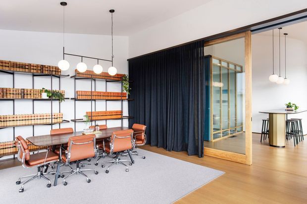 A bright contemporary office and meeting space with a long timber conference table surrounded by orange leather swivel chairs. Black open shelving displays rows of materials or books along the left wall, while a dramatic dark curtain and timber-framed glazed partition separate the meeting area from an adjacent kitchenette or breakout space. Pendant globe lights, pale timber flooring, and light walls create a refined, warm workplace interior.