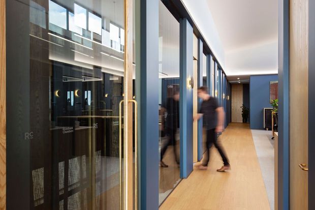 A contemporary commercial interior hallway with a warm timber floor, blue-painted structural and door frames, and a series of glass-fronted rooms or offices along both sides. The corridor has a clean linear layout, recessed ceiling lighting, and a glazed entry door with a brass-toned pull handle. Blurred figures indicate movement through the space, while reflected interiors are visible in the glazing.