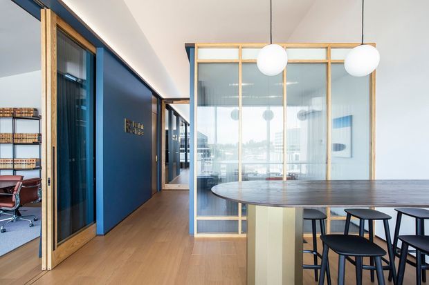 Contemporary commercial interior featuring an open dining or breakout area with a large dark timber island table and black stools. A blue feature wall, light timber-framed glazed and ribbed-glass partitions, timber flooring, and spherical pendant lights define the space. An office workspace is visible through the open doorway at left, while bright glazing and reflected furnishings extend the view into the background.