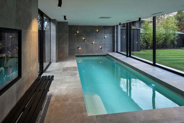 A sleek indoor swimming pool in a contemporary pavilion, with large frameless-looking glazed walls opening views to a landscaped lawn. The pool is surrounded by large-format grey stone or concrete-look tiles, dark charcoal wall surfaces, integrated wall lighting, a long dark timber bench, and a recessed fireplace or display niche. The space has a low flat ceiling, minimalist detailing, and a calm resort-like atmosphere.