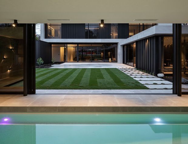 View from a covered interior terrace or poolside area across a rectangular swimming pool toward a landscaped internal courtyard. The courtyard features striped lawn, large pale stepping stones, dark vertical cladding, extensive floor-to-ceiling glazing, and warm architectural lighting. The composition emphasizes a contemporary indoor-outdoor connection and strong geometric lines.