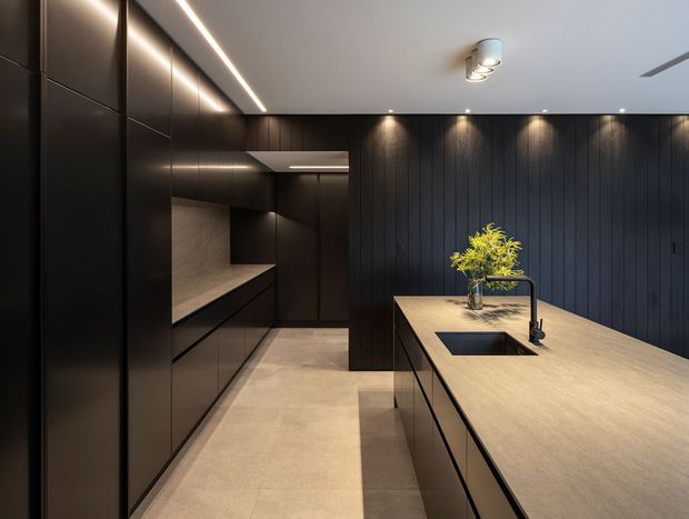 A sophisticated contemporary kitchen with a dark, streamlined palette. Floor-to-ceiling matte black cabinetry lines the left wall, complemented by dark vertical wall panelling at the rear. A large pale stone island with an integrated dark sink and black tap occupies the foreground. Recessed spotlights and linear LED lighting provide warm, focused illumination, while a small vase of greenery adds a natural accent.