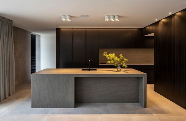 A sophisticated contemporary kitchen dominated by a large monolithic island with a dark grey textured finish and integrated sink. Full-height dark timber or dark-stained cabinetry lines the rear and right walls, with a warm wood-toned splashback and concealed lighting. Neutral stone or concrete-look flooring, minimal pendant-free ceiling lighting, and a restrained palette create a refined, moody atmosphere.