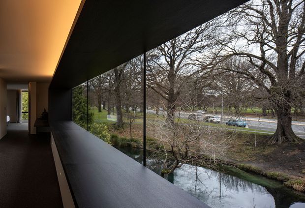 A contemporary interior corridor or landing with a long, dark built-in ledge running beside full-height glazing. The glazing overlooks a tree-lined waterway and road, while a warm concealed ceiling light and dark timber-toned finishes create a refined, moody atmosphere. A small seating or living area is visible at the far end.