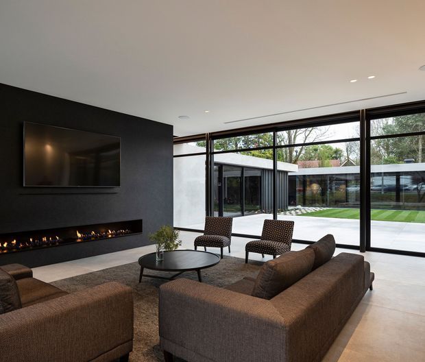Contemporary living room with a large brown sectional seating arrangement, two patterned lounge chairs, and a round dark coffee table. A black feature wall incorporates a wall-mounted television and long linear fireplace. Full-height black-framed glazing opens the room visually to a landscaped courtyard and adjacent modern pavilion. The space has a clean, refined fit-out with polished light flooring, recessed ceiling lighting, and strong indoor-outdoor connection.