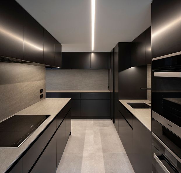 A sleek, narrow galley kitchen with full-height matte black cabinetry, pale stone-look benchtops and matching wall splashback, integrated black appliances, an induction cooktop, sink, and a linear recessed ceiling light. The restrained palette and handleless joinery create a dark, refined contemporary interior.