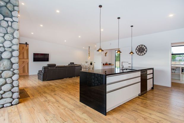 Open-plan kitchen and living area with a large central island, white flat-panel cabinetry, dark stone-look end panels and benchtops, pendant lighting, timber flooring, and a dark leather sofa. A distinctive rounded-stone feature wall and timber sliding barn-style door frame the living zone, while daylight enters through the windows.