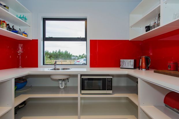 Bright compact kitchen with white open shelving and cabinetry, glossy red splashback panels, pale benchtops, stainless steel sink, and integrated microwave. A central window brings in natural light, while small appliances and dishware sit on the counters and shelves.