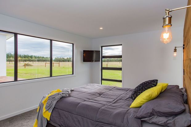 A bright contemporary bedroom with a low-profile bed dressed in charcoal-grey bedding and vivid yellow cushions and throw. Large black-framed windows and a glazed door provide views across open rural fields, while a timber feature wall and exposed industrial-style pendant and wall lights add warmth and character.