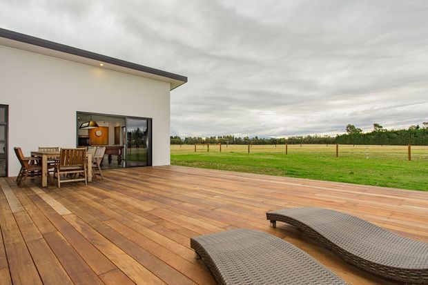 A contemporary single-storey home opens onto a broad timber deck overlooking a flat rural landscape. The deck features an outdoor dining setting and two curved woven sun loungers, while large glazed doors connect to the interior. The simple white rendered facade has dark trim and a shallow roof edge under an overcast sky.