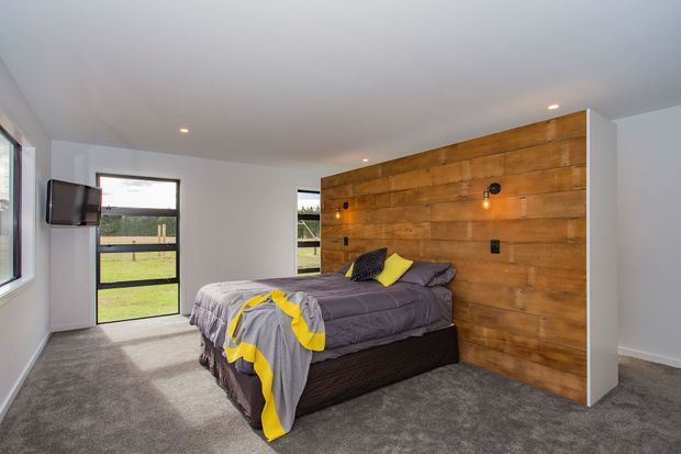 A contemporary bedroom with a double bed, grey carpet, white walls and ceiling, and a full-height warm timber feature wall behind the bed. The room has recessed ceiling lights, black-framed windows and a glass door opening to a grassy outdoor area. Grey and yellow bedding adds a bright accent.