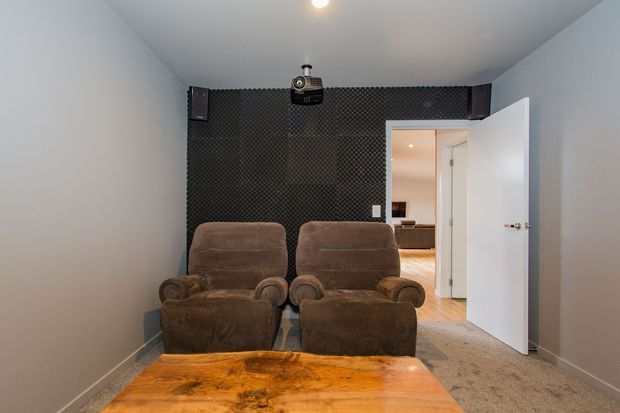 A compact private home theatre with two plush brown reclining cinema chairs facing a wall-mounted projector and speakers on a black acoustic-panel feature wall. A rustic timber table occupies the foreground, with grey carpet, pale walls, and an open door leading to a brighter adjoining room.